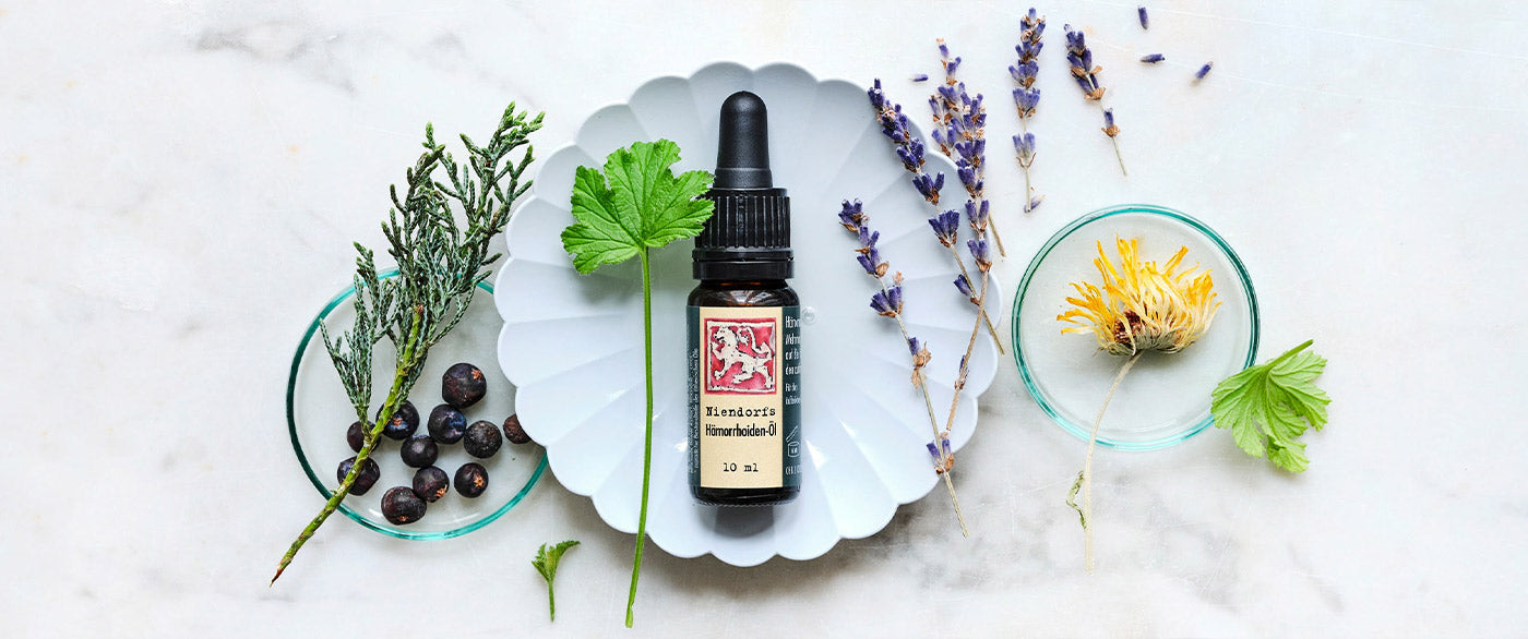 läschchen Hämorrhoiden-Öl umgeben von Wacholderbeeren, Lavendel und Kräutern