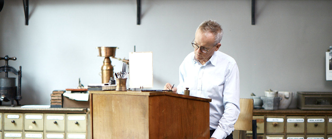 Mann schreibt an historischem Stehpult mit alten Apothekerschränken