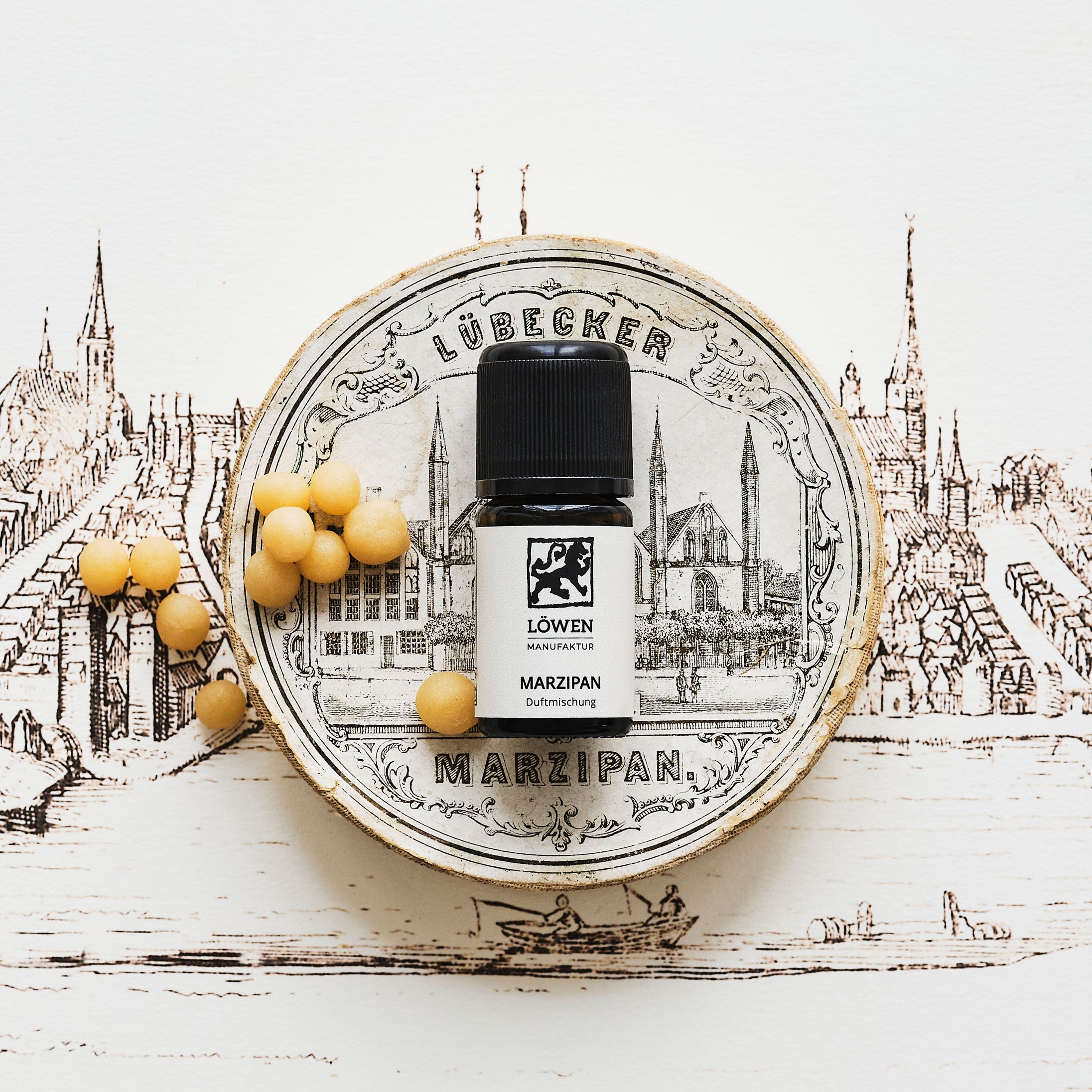 Duftmischung „Marzipan“ von Löwen-Manufaktur in Braunglasflasche mit schwarzem Deckel, inszeniert auf einer historischen Lübeck-Illustration mit Marzipanpralinen und nostalgischem Dosenmotiv.