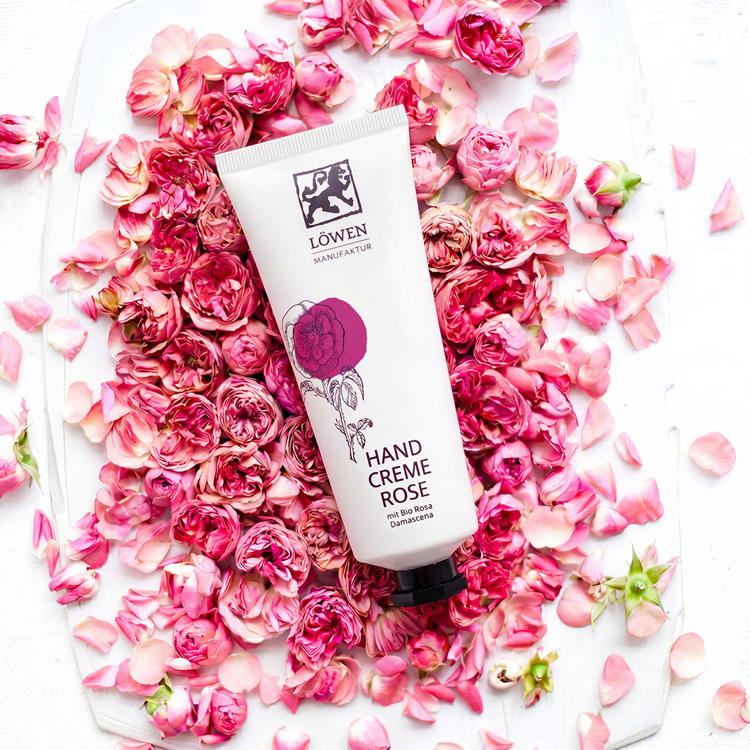 Handcreme Rose mit Bio Rosa Damascena, arrangiert mit rosa Rosenblüten und Blütenblättern als stimmungsvolle Produktpräsentation.