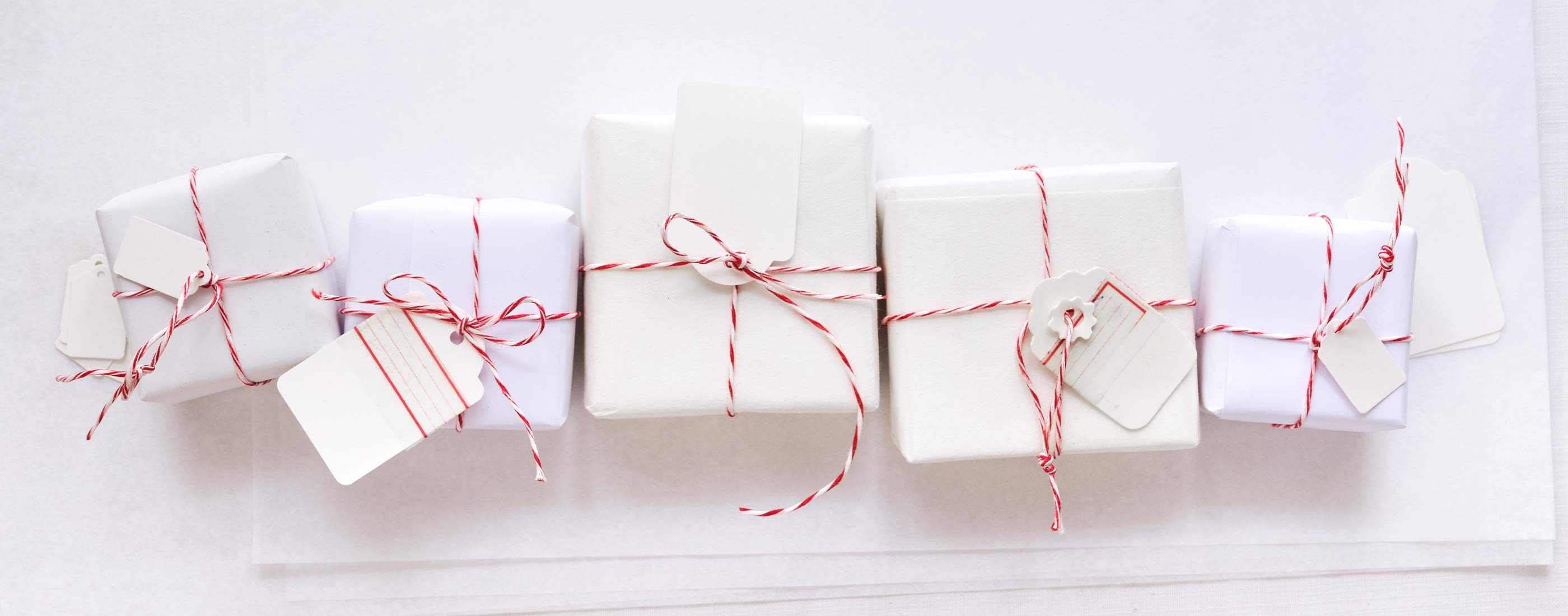 Fünf liebevoll verpackte Geschenke mit weißen Anhängern und rot-weißem Garn auf hellem Untergrund.