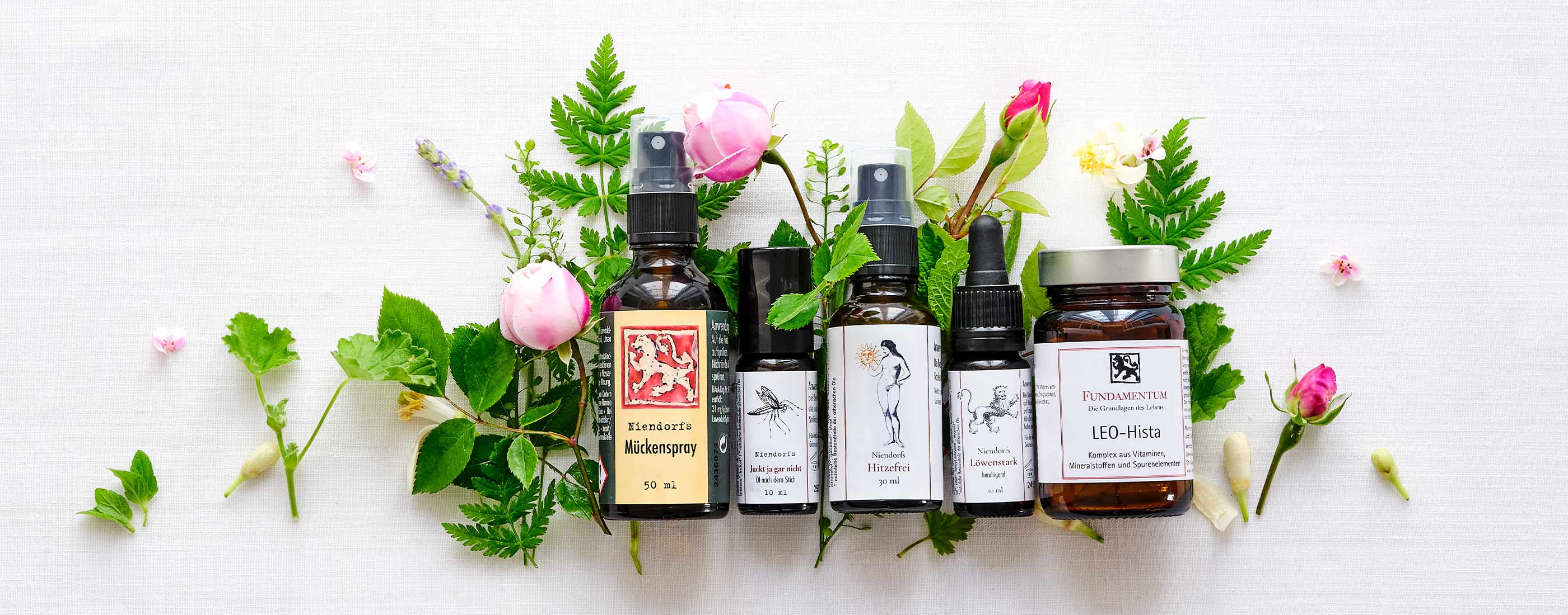 Sommerlieblinge der Löwen‑Manufactur mit Mückenspray, Roll‑On Juckt ja gar nicht, Hitzefrei Spray, Löwenstark Öl und LEO‑Hista Kapseln, dekorativ mit Blüten und Kräutern auf Leinentuch arrangiert