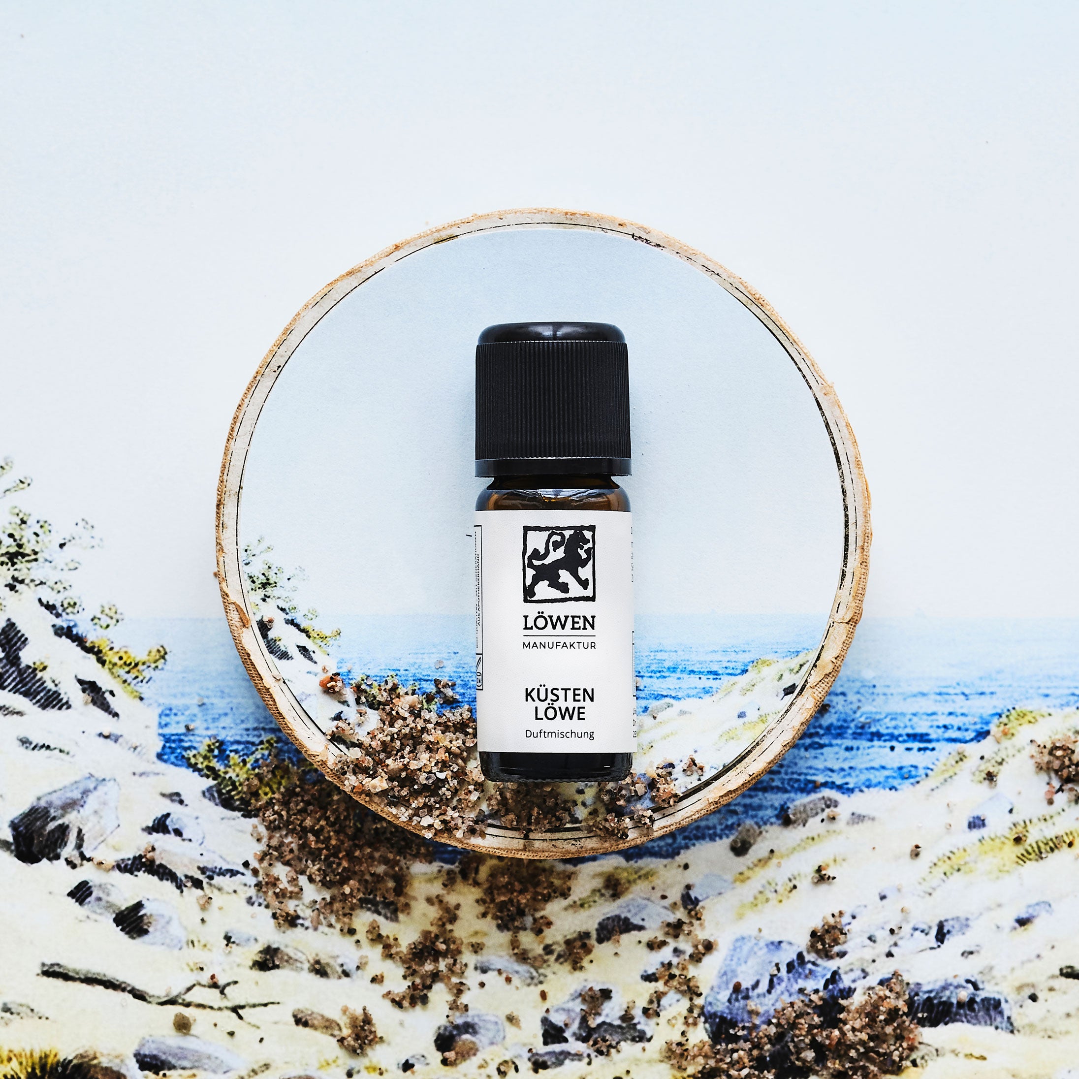 Duftmischung „Küsten Löwe“ von Löwen-Manufaktur in Braunglasflasche mit schwarzem Deckel, inszeniert auf einer maritimen Illustration mit Dünenlandschaft, Sand und Blick aufs Meer – umrahmt von einem Baumstammkreis.