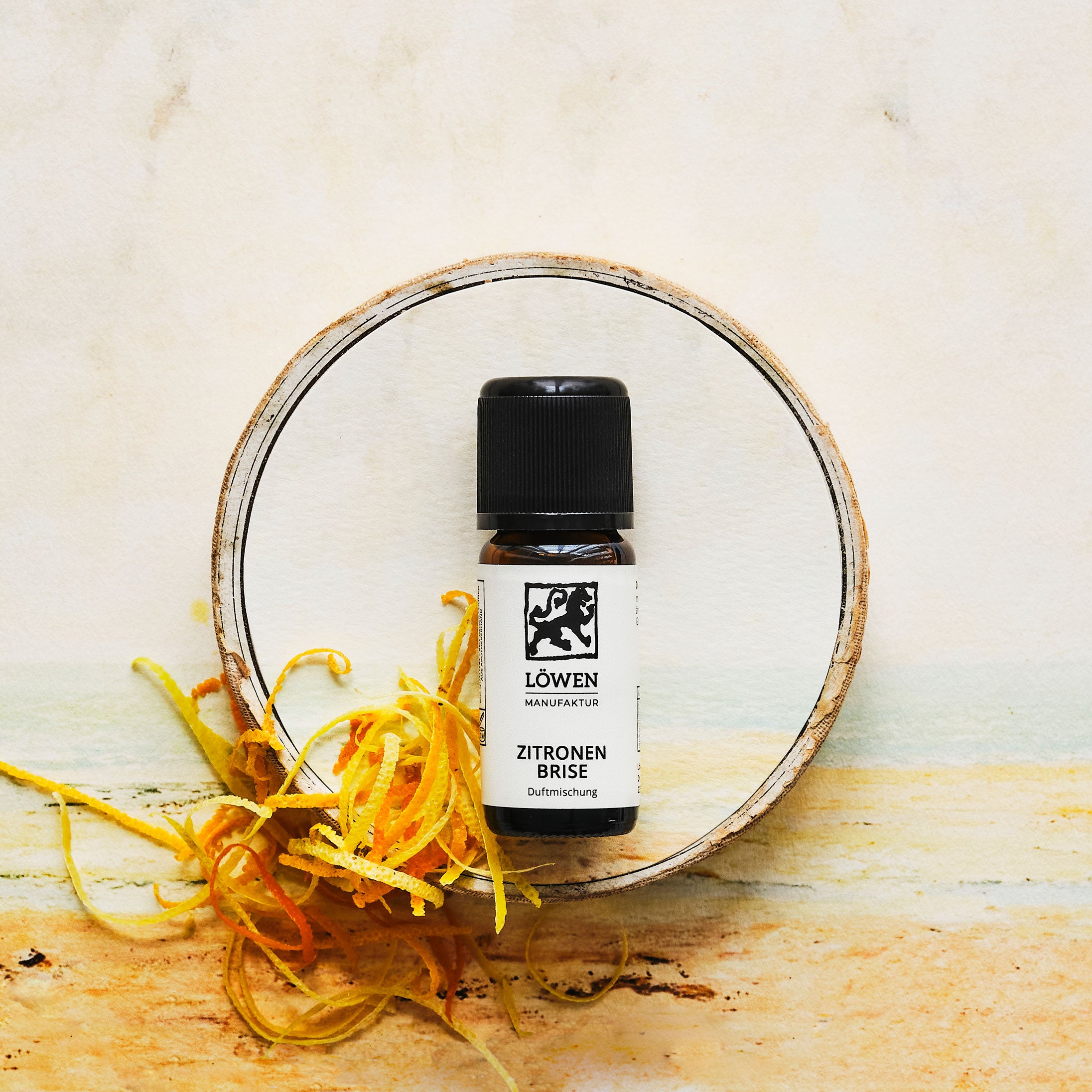 Duftmischung „Zitronen Brise“ von Löwen-Manufaktur in Braunglasflasche mit schwarzem Deckel, inszeniert auf einer sommerlich hellen Illustration mit Zitronenzesten und pastellfarbener Strandoptik, umrahmt von einem Baumstammkreis.