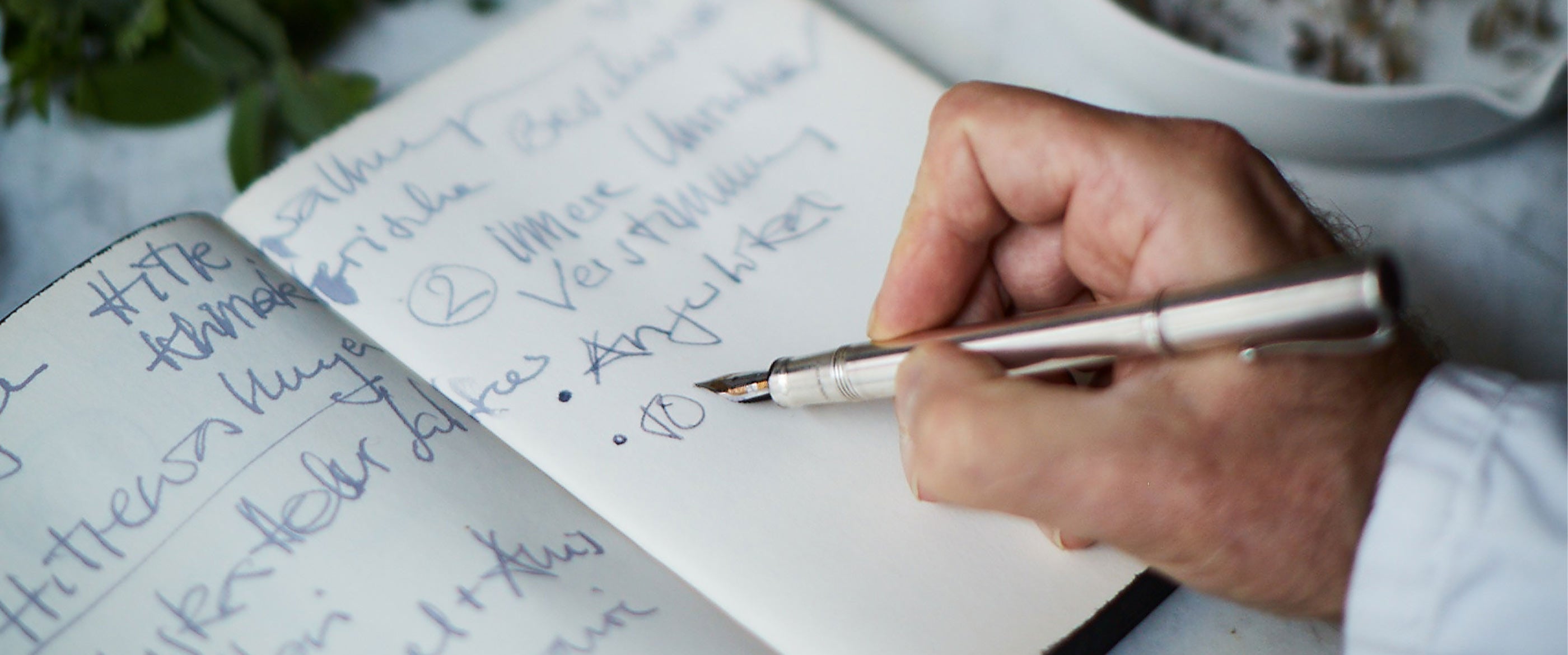 Eine Hand schreibt mit einem Füller in ein Notizbuch mit Pflanzen und Blüten daneben, symbolisch für kreative Natur-Rezepte oder Notizen.